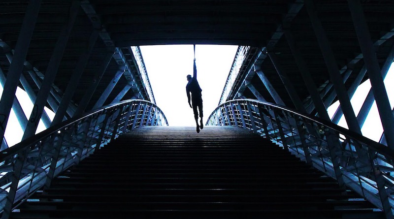 Erwan Tarlet hangs by one arm on a strap under a bridge