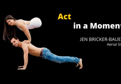 A female acrobat with no legs in a white leotard does a handstand arm press on top of another acrobat's back. He is shirtless and doing a plank