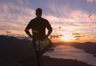 A man is silhouetted with his arms akimbo, sitting on the top of a pole, on the top of a mountain, overlooking a lake below, aglow in oranges and yellows of sunset