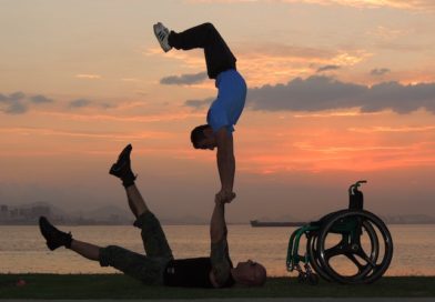 duo Dupla Mao Na Roda performing hand balancing by a lake at sunset. Photo description: There are bluish grey clouds and the sky is orange.Two handbalancers are silhoutted in the forefront. One hand balancer lays on the ground holding hands with the second hand balancer who is doing a handstand on the hands of the first. Also in the silhouette is the wheelchair of one of the handbalancers.