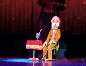 A puppet depicting an older man with a large comical mustache in a tweed suit with a cane sitting by a small table with a drink