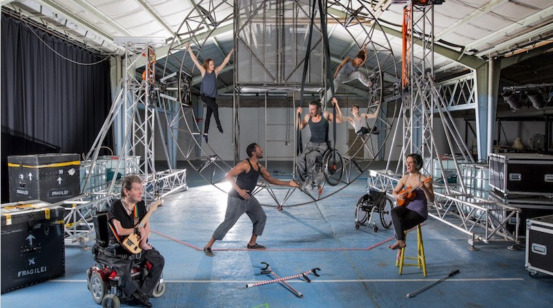 Seven performers disabled and non disabled surround a steel circular structure that they hang from . Two artists seated in front  play stringed unique instruments. Crutches lay on the floor . An artist who is a wheelchair user is suspended in his wheelchair from the structure.