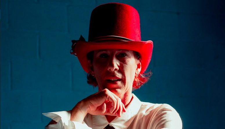 Aloysia Gavre staring into the camera thoughtfully, resting a hand on her chin, wearing a white button down and a large red hat