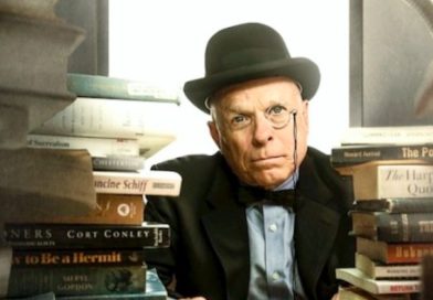 Jim Moore stares into the camera wearing spectacles and a black hat, surrounded by books