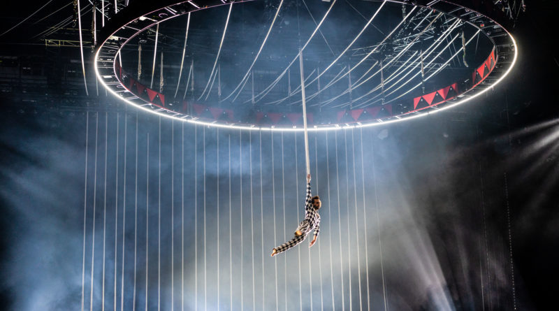an aerialist flies beneath circular scaffolding with a back drop of ropes