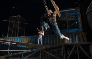 Two acrobats, one hanging above the stage, and one climbing a crate in the background