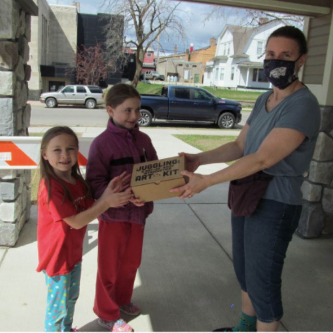 Outside an Arrowhead library, a young man hands a juggling kit to two girls wearing red