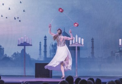 Lena Koehn (Köhn), German diabolo artist, performs tricks onstage for a crowd. She wears a long, white dress and faces a blue backdrop