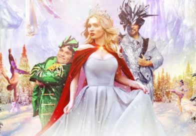 In Cirque Musica's 2022 holiday show, a female circus performer in a white gown and red cloak stands between a child performer in a Christmas tree costume and a man in a silver and brown costume with an antlered mask