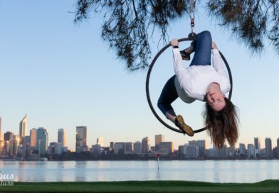 Circus artist and author Fleur Van Rens, Ph.D, hangs upside-down in her Lyra, suspended from a tree near Sydney Harbour. The skyline is visible behind her