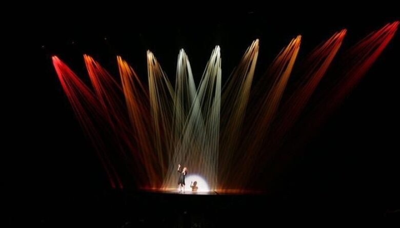 A single performer in center stage, illuminated under ten spotlights; the outer eight are red lights.