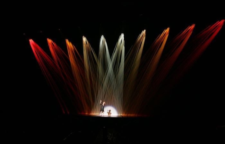 A single performer in center stage, illuminated under ten spotlights; the outer eight are red lights.