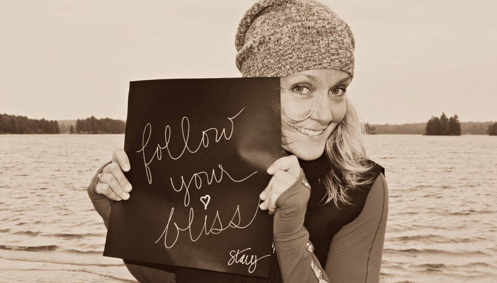 Circus consultant and CircusTalk CEO Stacy Clark, a smiling blonde Canadian woman wearing a beanie, holds a sign with "follow your bliss" written in cursive while standing on the shores of a local river