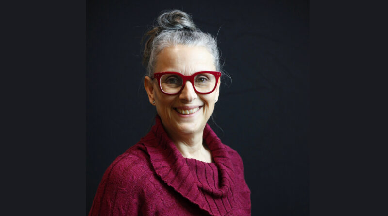 Nadia Drouin, Executive Director of En Piste, is a middle-aged woman with gray hair in a bun, a smile, and smart attire. She wears a red cowl-necked sweater that matches her red-framed glasses.