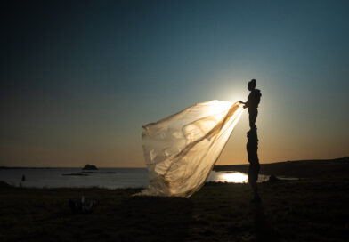 Circus Barcode artist Alexandra Royer in performance on a beach, part of her work with "Elementerre". She stands in profile silhouette with one side to the sunset, unfurling a billowing plastic sheet with both hands